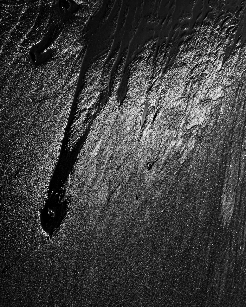 黒い砂浜に水が流れ落ちる抽象的なモノクロ写真。光と影のコントラストが強く、流れるような線と粒状の質感が強調されたクローズアップ。