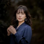 A portrait of a woman wearing a navy blouse and pearl earrings, bathed in soft light against a dark background, posing with her hands lightly clasped in front of her chest.