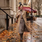 秋のパリの街並みを歩く、ベージュのトレンチコートとグレーのニットワンピースを着た笑顔の女性。濡れた石畳に落ち葉が散り、背景にカフェの赤い日除けが見える